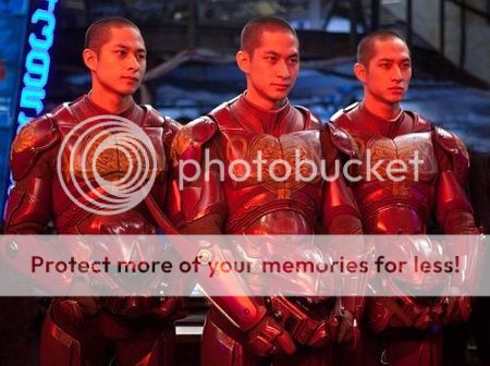 The Wei triplets standing together in their red flight suits, ready for deployment in their giant three-armed robot