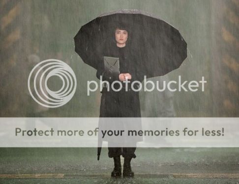Mako Mori all in black and wearing black boots, under an umbrella in the rain